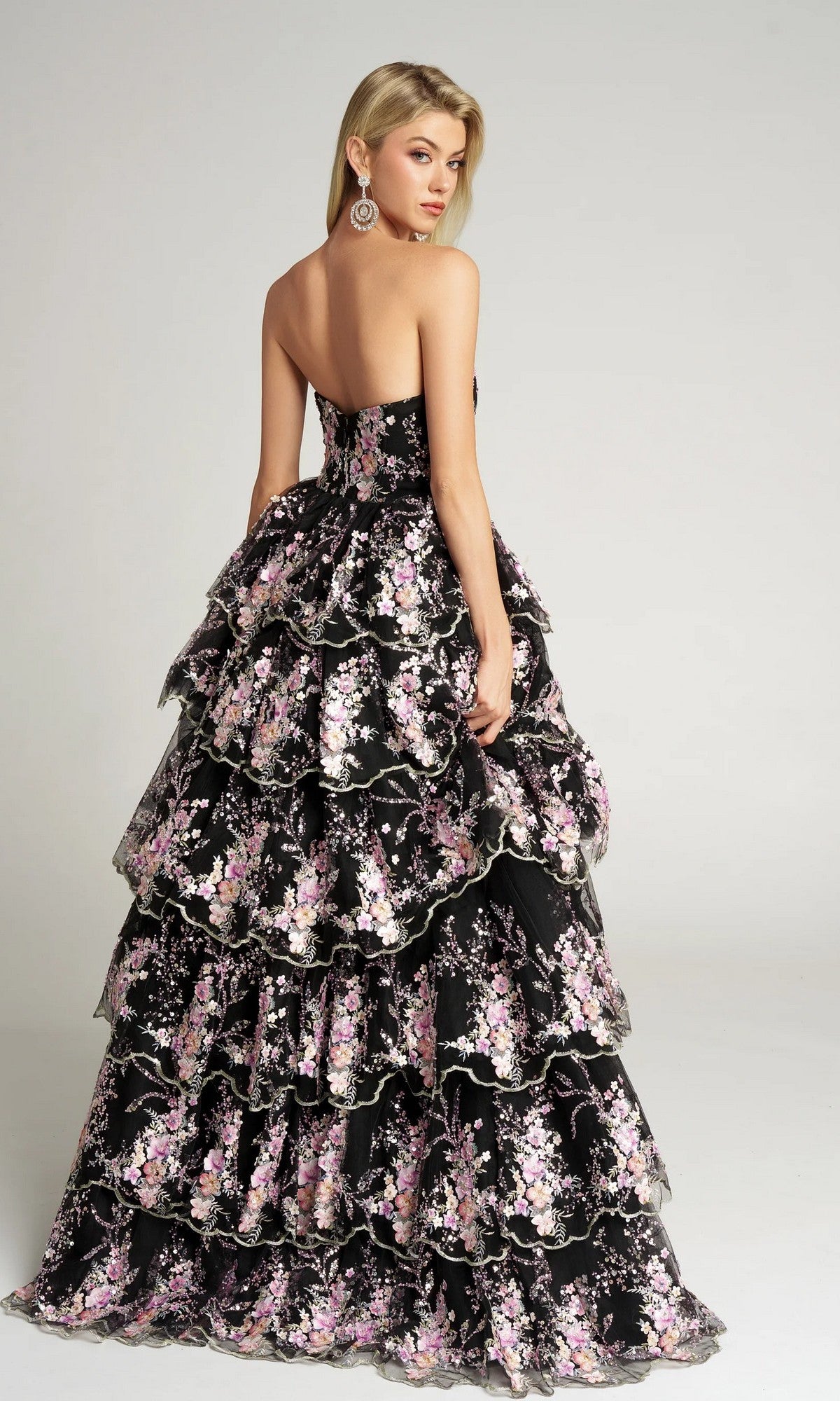 A woman in the Portia and Scarlett PS26051 Strapless Floral Ball Gown, featuring pink and white embroidered floral appliqué, stands facing away while looking over her shoulder.